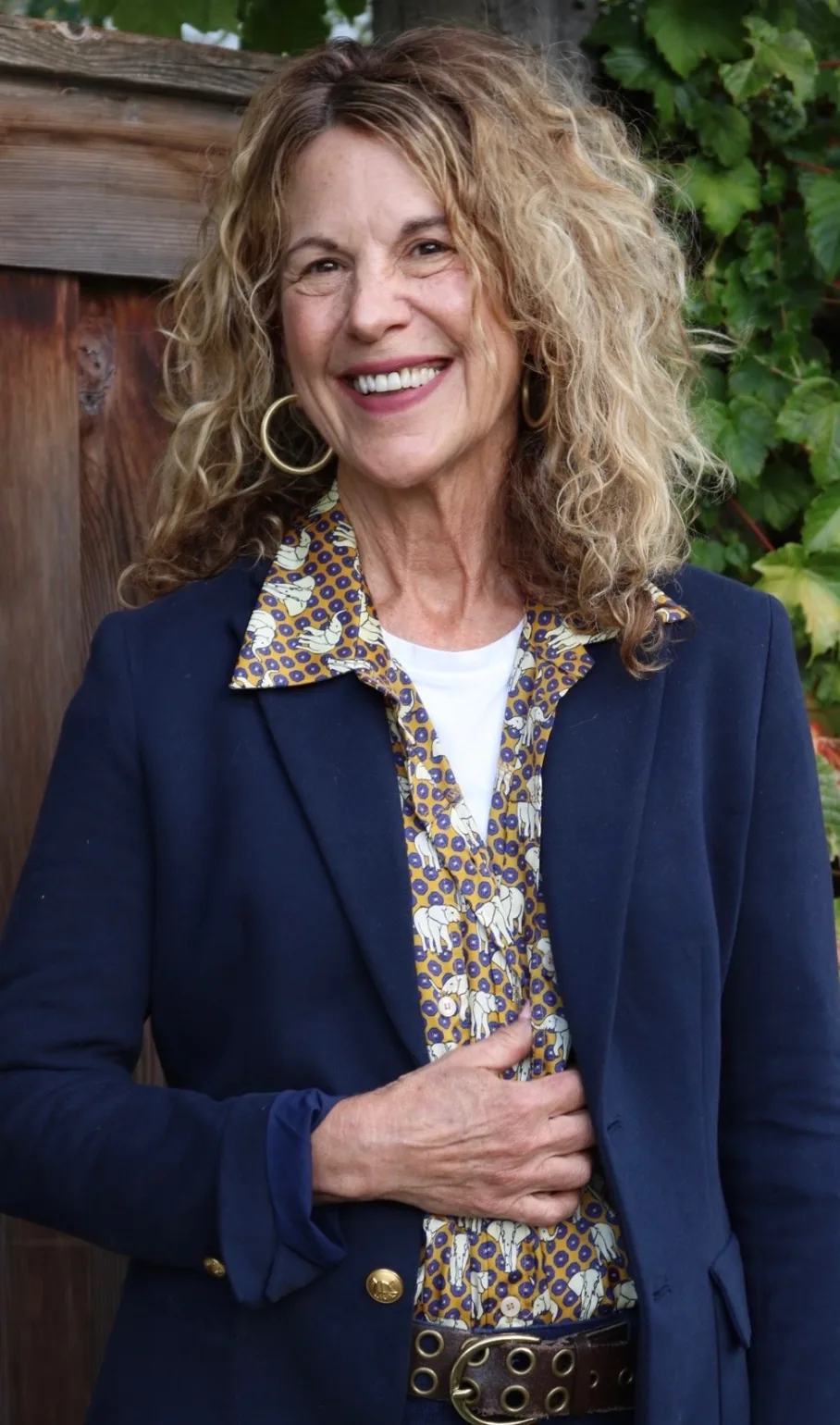 Sharon Davis, smiling, hands resting on her middle, outdoors against a weathered fence and ivy.